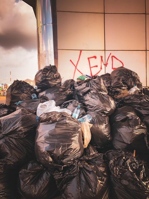 A large pile of black plastic rubbish bags is stacked against a modern building with a reflective metal facade, partially covered by graffiti spray-painted in red that reads 'XEND'. The bags are filled with waste and appear to be tightly knotted at their tops, with some slightly torn or bulging. A few discarded plastic water bottles and paper or plastic packaging are visible protruding from the bags, indicating mixed waste. The background shows a cloudy sky with a hint of sunlight creating a soft glow on the metallic surface of the building. The scene depicts an outdoor urban setting, likely an area awaiting collection as part of private waste management or rubbish clearance services, aligning with the concept of alternative disposal methods outside regular council collection schedules. The overall environment suggests the importance of professional rubbish removal, such as that provided by Rubbish Removal Hounslow, to manage accumulation and maintain cleanliness in a city setting.