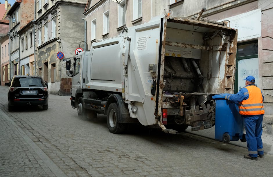 A street scene shows a white refuse collection vehicle with an open rear compartment, positioned on a cobblestone road next to a row of multi-storey buildings with aged facades. The vehicle is equipped with a hydraulic lift system and has visible rust spots and dirt on its metal surfaces, indicating use. A person wearing a blue uniform, orange high-visibility vest, and a blue cap is standing beside the vehicle, emptying a blue wheeled bin into the rear chamber. The surrounding environment features another parked car, pedestrian signage, and a narrow sidewalk, with muted natural lighting suggesting overcast weather. The scene reflects a typical urban waste collection process, representing an alternative to scheduled rubbish removal services by highlighting on-site waste disposal activities managed privately, as performed by companies like Rubbish Removal Hounslow.