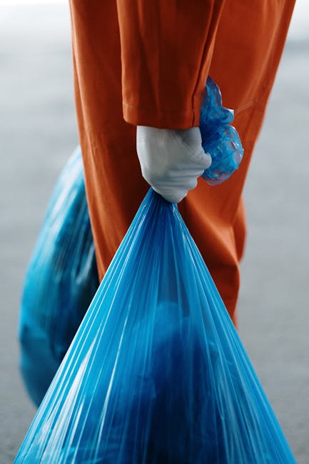 A person wearing orange protective trousers and white gloves is holding a large blue plastic bag filled with waste. The person's hand grips the bag's tied section, which is secured with a knot. The background appears to be an outdoor setting with a blurred, neutral-toned surface, possibly a paved area or pavement. The blue trash bag is semi-transparent, revealing some visible contents inside, and has a smooth surface with slight glossiness. The orange trousers provide a stark contrast to the blue bag, emphasizing the manual handling of rubbish. The scene captures an element of waste collection or disposal activity, which could be part of a private rubbish removal service, reflecting an on-site clearance operation. The overall environment seems well-lit, with natural lighting that highlights the textures of the bag and the clothing. This image visually supports themes of waste management, independent refuse collection, and waste handling operations, as seen in the context of rubbish removal services offered by companies like Rubbish Removal Hounslow.