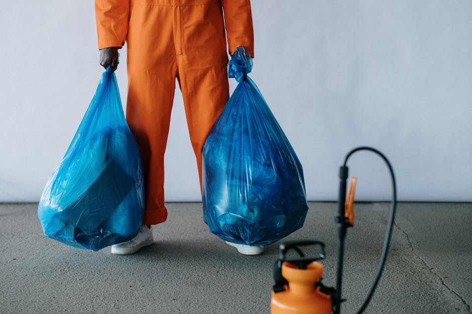 A person dressed in an orange protective jumpsuit and white footwear is holding two large blue plastic rubbish bags, one in each hand, standing on a gray concrete floor against a plain light gray wall. The bags are filled with waste material and appear slightly compressed and semi-transparent, revealing some contents inside. In the foreground, there is a partially visible orange and black industrial waste sprayer or blower, with a flexible hose extending towards it, suggesting equipment used in waste collection or site clearing. The environment appears to be an indoor or controlled setting suitable for waste management activities, with no other objects or background features visible. This scene relates to rubbish removal services, highlighting the process of handling and preparing waste for disposal or transportation, consistent with services offered by companies such as Rubbish Removal Hounslow, which specialise in private and on-site waste clearance solutions, including collection of domestic, commercial, or construction debris.