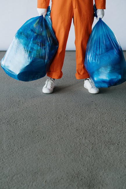 A person wearing orange protective trousers and white gloves is holding a large blue plastic bag filled with waste. The person's hand grips the bag's tied section, which is secured with a knot. The background appears to be an outdoor setting with a blurred, neutral-toned surface, possibly a paved area or pavement. The blue trash bag is semi-transparent, revealing some visible contents inside, and has a smooth surface with slight glossiness. The orange trousers provide a stark contrast to the blue bag, emphasizing the manual handling of rubbish. The scene captures an element of waste collection or disposal activity, which could be part of a private rubbish removal service, reflecting an on-site clearance operation. The overall environment seems well-lit, with natural lighting that highlights the textures of the bag and the clothing. This image visually supports themes of waste management, independent refuse collection, and waste handling operations, as seen in the context of rubbish removal services offered by companies like Rubbish Removal Hounslow.