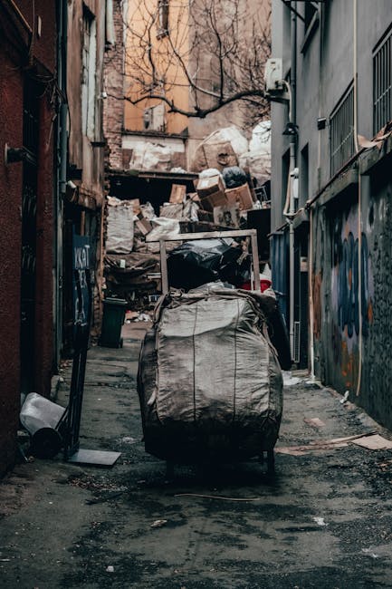 A narrow urban alleyway cluttered with numerous piles of waste and discarded items, including large black and white garbage bags, cardboard boxes, and loose trash. The alley is bordered by weathered brick and concrete walls, one side featuring graffiti and the other with metal railings and utility pipes. In the background, there is an accumulation of waste materials stacked against a makeshift wooden barrier, with leafless tree branches extending overhead, indicating late autumn or winter. The ground surface is uneven, dirty, and stained, with scattered debris, while a pushcart covered with a large white tarpaulin is positioned in the center foreground, suggesting the presence of waste collection or removal activity. The scene reflects an environment where independent rubbish disposal or collection services, such as those provided by Rubbish Removal Hounslow, might operate to manage large amounts of refuse from urban areas.