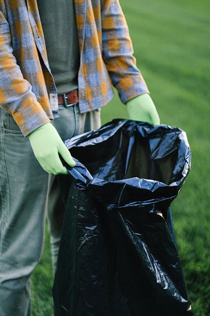 A worker in a blue uniform and orange high-visibility vest is standing next to a blue rubbish bin, momentarily releasing it into the rear compartment of a white waste collection truck with a rusted, open loading area. The truck is parked on a narrow, cobbled street lined with older, multi-storey buildings featuring a mix of beige and gray facades, some showing signs of weathering and patchwork repairs. The environment appears urban, with a black parked vehicle visible on the left side of the street and a no parking sign mounted on a pole further along. The scene is illuminated by natural daylight, with an overcast sky possibly diffusing the light evenly across the area. This image exemplifies a private rubbish collection service, likely undertaken by Rubbish Removal Hounslow, supporting alternative waste handling outside local authority disposal systems, with the worker engaged in on-site waste removal or commercial rubbish collection efforts using specialized equipment designed for urban refuse management.