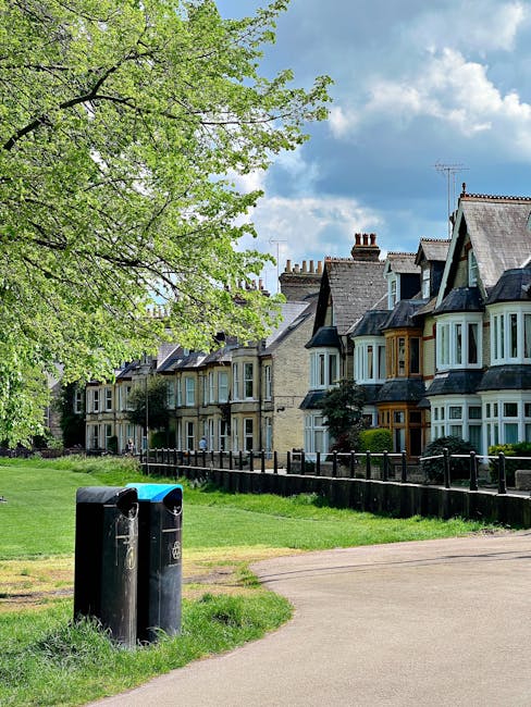 In a residential area with a row of traditional terraced houses featuring stone and brick facades, multi-pane bay windows, and sloped slate roofs, a large leafy tree with bright green foliage extends its branches over the scene in the foreground. The sky above is partly cloudy with patches of blue visible through the cloud cover. In front of the houses, a neatly maintained grassy verge runs alongside a paved walkway, with two black plastic waste bins positioned near the edge of the path. The bins are cylindrical, with one slightly taller than the other, and appear to be used for general waste disposal. A black metal fence separates the grassy area from the pavement, providing a boundary between the footpath and the private gardens of the properties. The scene depicts a calm, well-kept neighborhood, illustrating the typical setting for private rubbish collection services such as those provided by Rubbish Removal Hounslow, which may include on-site clearance or alternative waste handling methods.