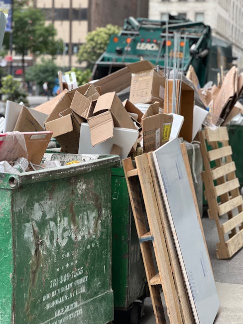 A large green waste skip overflowing with various types of rubbish and packaging materials, including multiple cardboard boxes, some flattened and others partially assembled, as well as wooden pallets and a few white polystyrene foam pieces. The skip, which appears weathered with visible dirt and scuff marks, is situated outdoors on a paved surface, likely in an urban environment. In the background, a refuse collection vehicle with visible branding and orange safety lights is part of the scene, alongside blurred trees and high-rise buildings, indicating a city street setting. The overall composition suggests a scene of commercial or domestic waste collection, possibly handled by private waste management services such as Rubbish Removal Hounslow, offering alternative options for bulky waste disposal outside of regular council collection schemes. The lighting is natural, typical of daytime conditions, emphasizing the rough textures and varied materials of the scattered dumping area in an urban environment focused on waste removal tasks.