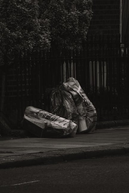 A large green waste skip overflowing with various types of rubbish and packaging materials, including multiple cardboard boxes, some flattened and others partially assembled, as well as wooden pallets and a few white polystyrene foam pieces. The skip, which appears weathered with visible dirt and scuff marks, is situated outdoors on a paved surface, likely in an urban environment. In the background, a refuse collection vehicle with visible branding and orange safety lights is part of the scene, alongside blurred trees and high-rise buildings, indicating a city street setting. The overall composition suggests a scene of commercial or domestic waste collection, possibly handled by private waste management services such as Rubbish Removal Hounslow, offering alternative options for bulky waste disposal outside of regular council collection schemes. The lighting is natural, typical of daytime conditions, emphasizing the rough textures and varied materials of the scattered dumping area in an urban environment focused on waste removal tasks.