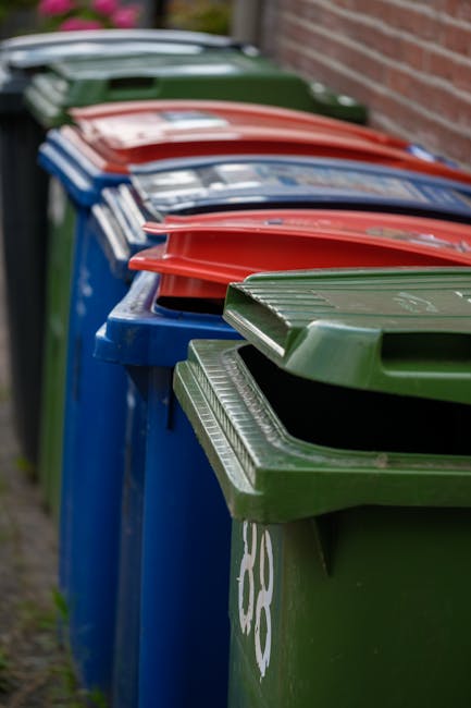 A row of large wheeled rubbish bins lined up along a pavement next to a brick wall, with the bins in various colors including green, blue, and red, each fitted with matching hinged lids. The green bin in the foreground is partially open, revealing a textured finish on its surface. The other bins are positioned behind it, with some lids slightly tilted or closed, and are arranged in a straight line extending into the background. The bins are situated outdoors, likely in a residential area, with natural daylight illuminating the scene. The environment appears clean and orderly, reflecting the typical setting for private waste collection or alternative rubbish removal services, such as those provided by Rubbish Removal Hounslow. The focus on the standard household waste containers suggests a context of domestic rubbish management rather than municipal collection.