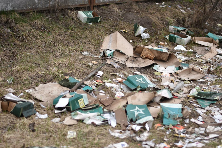 The image shows a patch of ground covered in various discarded cardboard boxes, some flattened and torn, with a predominantly green and white color scheme, and a visible star logo on several boxes. The boxes are scattered across a grassy area adjacent to a metal fence, with some leaning against the fence and others lying flat on uneven soil mixed with patches of grass. Among the debris, there are small pieces of paper and plastic wrapping, along with a long, dark pipe or rod slightly bent and partially buried under the waste. The environment appears to be an outdoor space, possibly a neglected or dumped refuse area, where rubbish removal services might be needed to clear the scattered packaging waste. The lighting appears natural, with diffuse daylight illuminating the scene, emphasizing the cluttered and unmanaged state of the refuse, which aligns with ideologies of alternative waste handling and private rubbish collection efforts by companies such as Rubbish Removal Hounslow.