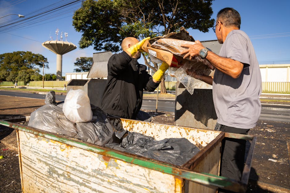 Two men are engaged in rubbish collection at an outdoor waste disposal site under clear blue skies. One man, dressed in a black jacket and yellow gloves, is lifting a cardboard box filled with waste materials from a large, open, rusted metal container. The other man, dressed in a light grey T-shirt and wearing glasses, is holding the box steady as the first man places it into the container. Surrounding the bin, there are several black and white plastic rubbish bags, some knotted and others loose, resting on the container's edge. In the background, there are trees with green foliage, a tall, grey water tower, and a fence, indicating a semi-urban area. The scene is well-lit by natural daylight, and the surface beneath the containers appears to be paved. This image exemplifies onsite waste handling, illustrating the process of manual rubbish removal, a service offered by companies like Rubbish Removal Hounslow as an alternative to private skip hire or council collection.