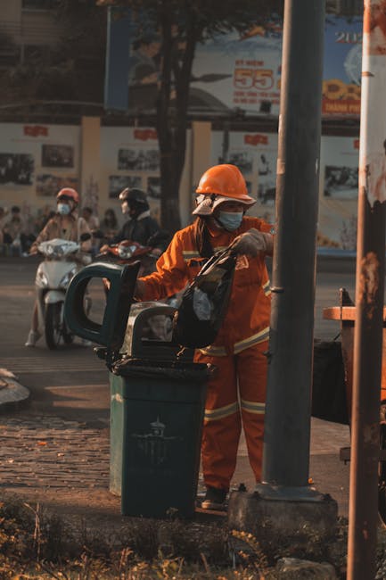 Two men are engaged in rubbish collection at an outdoor waste disposal site under clear blue skies. One man, dressed in a black jacket and yellow gloves, is lifting a cardboard box filled with waste materials from a large, open, rusted metal container. The other man, dressed in a light grey T-shirt and wearing glasses, is holding the box steady as the first man places it into the container. Surrounding the bin, there are several black and white plastic rubbish bags, some knotted and others loose, resting on the container's edge. In the background, there are trees with green foliage, a tall, grey water tower, and a fence, indicating a semi-urban area. The scene is well-lit by natural daylight, and the surface beneath the containers appears to be paved. This image exemplifies onsite waste handling, illustrating the process of manual rubbish removal, a service offered by companies like Rubbish Removal Hounslow as an alternative to private skip hire or council collection.
