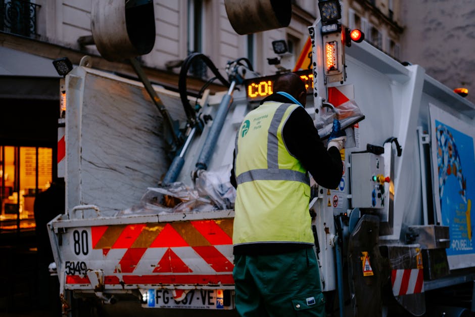 A waste collection operator dressed in a black jacket and high-visibility yellow vest with reflective strips is operating a modern rubbish collection vehicle on a city street during dusk. The vehicle, which is primarily white with red and orange reflective markings, is equipped with hydraulic arms and electronic controls visible on the side. The rear of the truck is open, revealing a load of mixed waste bags and debris, some of which are crumpled paper or plastic, piled inside the container. The operator is focused on the control panel, possibly updating data or confirming collection details, with a background showing blurred building windows with warm lighting, indicating an urban environment. The scene highlights the practical aspects of private waste handling, such as on-site collection and transportation, reflecting the services offered by rubbishremovalhounslow.org.uk. The lighting is natural and subdued, emphasizing the work activity during early evening hours, and the environment suggests a routine waste collection operation in a residential or commercial area.