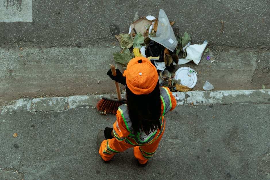 A municipal worker dressed in bright orange high-visibility clothing and a matching orange cap is seen from above, standing on a grey pavement. The worker is holding a broom with a wooden handle and black bristles, sweeping an area of street debris and rubbish. In front of the worker, there is a small pile of mixed waste including crumpled paper, leaves, and a broken plastic container, with some scattered on the curb. The pavement shows signs of wear, with visible cracks and darker stains, and a curb separates the street from the sidewalk. The background includes a section of asphalt roadway with a white line marking, and the scene is evenly illuminated, suggesting daytime natural light. This image relates to private waste collection services such as those provided by Rubbish Removal Hounslow, emphasizing street cleaning and rubbish clearance activities undertaken by trained waste management professionals as part of alternative rubbish removal solutions.