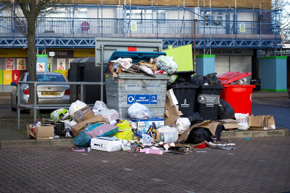 A large collection of mixed household and commercial rubbish is piled on the pavement in front of a set of recycling containers and a parked silver car. The rubbish includes black, green, and red plastic waste bins, along with open and closed cardboard boxes, crumpled paper, plastic bags, and assorted packaging materials. Some items are spilling onto the surrounding brick-paved area, while the open middle container is overflowing, with waste such as paper, cardboard, and plastic visible inside. In the background, a building with a blue construction fence and signage indicating a fish bar can be seen, with storefronts and shops bordering the scene. The overall setting appears to be in an urban area, with the waste accumulated temporarily outside, possibly awaiting collection or disposal by a private rubbish removal service like Rubbish Removal Hounslow.