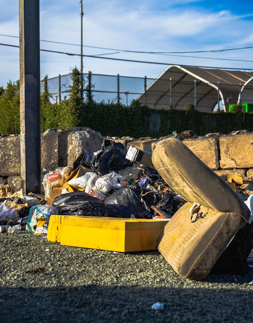 A close-up view of an outdoor rubbish pile situated on a gravel surface, featuring a variety of waste materials including black plastic garbage bags, a yellow plastic container, a discarded worn-out car seat upholstery with visible fabric and foam padding, and broken pieces of wood or debris. The background shows a stone wall composed of large, irregularly shaped beige and light brown stones, with some greenery such as small bushes and trees behind it. A metal utility pole flags the scene, with overhead power lines running across the sky, which is partly cloudy with patches of blue indicating daytime. To the right, there is a glimpse of a large, curved, metallic structure that appears to be a shed or canopy, and additional details like the nearby pavement edge suggest this waste is located on a roadside or parking area. The scene reflects typical elements involved in independent waste handling or rubbish removal services, as offered by companies like Rubbish Removal Hounslow, showcasing the need for proper disposal and clearing of unwanted debris.