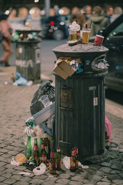 A large collection of mixed household and commercial rubbish is piled on the pavement in front of a set of recycling containers and a parked silver car. The rubbish includes black, green, and red plastic waste bins, along with open and closed cardboard boxes, crumpled paper, plastic bags, and assorted packaging materials. Some items are spilling onto the surrounding brick-paved area, while the open middle container is overflowing, with waste such as paper, cardboard, and plastic visible inside. In the background, a building with a blue construction fence and signage indicating a fish bar can be seen, with storefronts and shops bordering the scene. The overall setting appears to be in an urban area, with the waste accumulated temporarily outside, possibly awaiting collection or disposal by a private rubbish removal service like Rubbish Removal Hounslow.