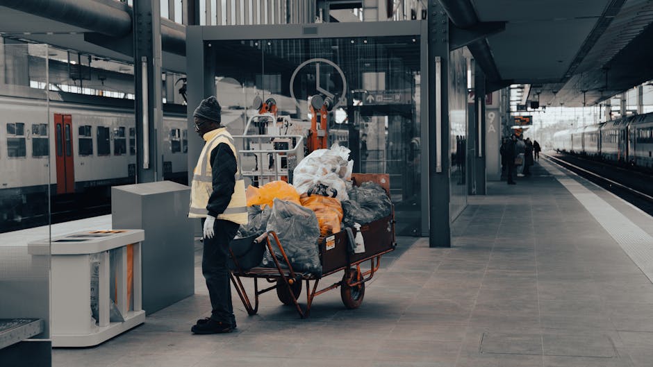At a modern railway station platform, a worker dressed in dark clothing, a beanie, and a high-visibility vest is standing next to a large wheelbarrow loaded with various types of rubbish, including black and orange plastic bags, cardboard, and other waste materials. The materials in the wheelbarrow display a mix of textures, with smooth plastic bags and crumpled cardboard surfaces, all piled haphazardly. Behind the worker, a glass enclosure with white metal framing reveals automated waste collection equipment, indicating an effort for efficient disposal or recycling processes. To the left, a small white waste bin with a partially open lid is placed on the platform, suggesting ongoing rubbish management as part of the station's cleaning routine. On the right side of the image, the platform extends with a slightly curved concrete surface, leading toward the train tracks where a train is parked, and a few figures are visible in the background, perhaps station staff or passengers. The environment is well-lit with natural daylight, and the overall scene emphasizes a clean, structured area dedicated to waste handling consistent with the practices of independent rubbish removal services such as Rubbish Removal Hounslow.