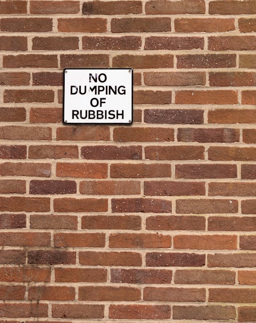 A rectangular white metal sign with black text reading 'NO DUMPING OF RUBBISH' is mounted on a red-brown brick wall. The brick wall features a traditional running bond pattern with a mix of slightly darker and lighter bricks, some showing subtle weathering or variations in texture. The wall surfaces are even with mortar joints between the bricks. The sign is positioned approximately one-third from the top left of the image, centered horizontally on the wall. The scene is lit evenly, without visible shadows or natural light sources, indicating outdoor ambient lighting, and the overall setting appears to be an exterior urban or residential area potentially associated with private waste management or rubbish clearance services, such as those provided by Rubbish Removal Hounslow, emphasizing the importance of maintaining designated refuse areas free from illegal dumping.